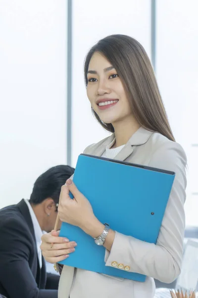 Beautiful Asian businesswoman standing near glass window with blue document folder in her hand. During her working hours. She smiled happily.