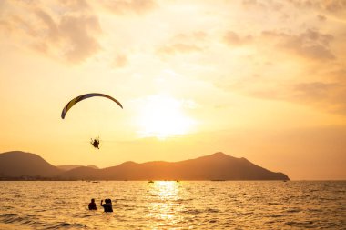 Paraşütçü, gölgeye karşı paraşütle atlarken güneş dağa ve denize batıyor. Spor turizmi yaz tatili konsepti.