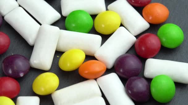 Γλυκά χρωματιστά καραμέλες skittles και μέντα μάσημα μαξιλάρια, χρωματιστά γλυκά περιστρέφονται σε ένα μαύρο πιάτο — Αρχείο Βίντεο