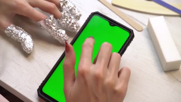 Una niña durante una manicura utiliza un teléfono inteligente con una pantalla verde. Servicio de manicura hecho a sí mismo. Manicurista pinta las uñas con esmalte de gel rosa. Uñas rojas cuidadas. Aplicación de esmalte de uñas. — Vídeos de Stock