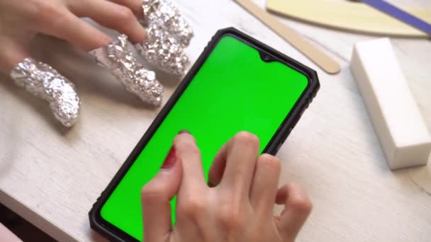 En flicka under en manikyr använder en smartphone med en grön skärm. Självgjord manikyrtjänst. Manicurist målar naglar med rosa gellack. Manikyrerade röda naglar. Nagellacksapplicering. — Stockvideo
