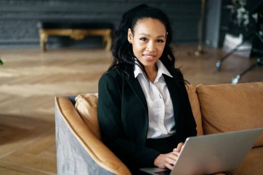 Çalışma alanındaki bir dizüstü bilgisayara mesaj yazarak çalışıyor. Ofiste resmi bir takım elbiseyle oturan kendine güvenen bir iş kadını afro görünümlü..
