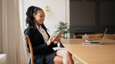 Ahşap bir masadaki ortak çalışma alanında dizüstü bilgisayara mesaj yazıyor. Ofiste resmi bir takım elbiseyle oturan kendine güvenen bir iş kadını afro görünümlü..