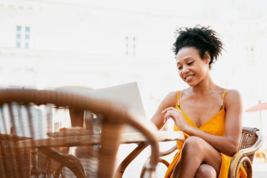 Bir kafede çalışırken, genç bir kadın gülümser ve dizüstü bilgisayarına bir mesaj yazar. Sarı ışıklı bir elbiseyle bir yaz restoranında oturmak. Afro-Amerikalı görünüşü ve siyah kıvırcık saçları var..