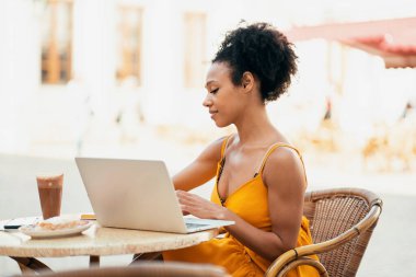 Restoranda serbest çalışan biri. Bir dizüstü bilgisayar ve tablet üzerinde çalışıyor, yeni bir tasarım üzerine online bir proje yapıyor. Afro-Amerikan görünümlü kendine güvenen bir kadın bir yaz kafesinde oturuyor..