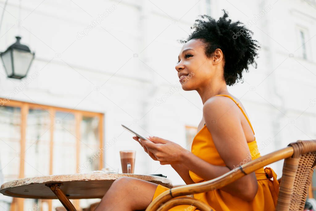 A student in a cafe on the street, a young woman working smiling typing a message on a tablet. African-American appearance and black curly hair. Sitting in a summer restaurant after a pandemic.