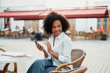 Afro görünümlü bir kadın elinde bir telefonla gülümsüyor ve bir yazlık kafede iş arkadaşına mesaj yazıyor..
