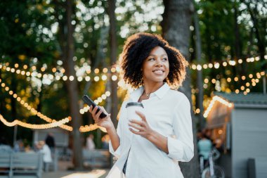 Elinde telefon ve kağıt bardakta sıcak kahve olan Afro görünümlü bir kadın parkta yürüyor. O dinleniyor ve sokakta zaman geçiriyor. Gülümser, dişlerini gösterir, rahat beyaz bir gömlek, kıvırcık saçlar..