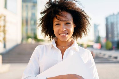Kameraya gülümseyen Afro görünümlü bir menajerin portresi. Bir öğrenci enstitünün yanında duruyor, kıvırcık saçlı ve beyaz iş gömlekli..