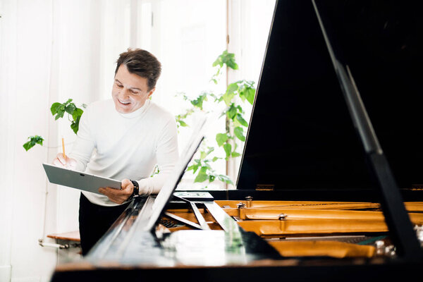 Professional activity of an adult. A male musician writes down the notes for a new song. The artist works at home and plays the piano. He teaches at a music school.