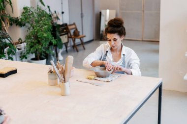 Profesyonel bir zanaatkar seramiklerden yemek yapar. Önlüklü bir kadın çömlek atölyesinde çalışıyor. Dekoratif ürün stüdyosunda üniformalı bir çalışan. Bir seramik porselenden bir tabak yapar..