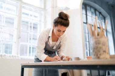 Stüdyoda çalışan bir kadın kilden bulaşık yapıyor. Dizayn için seramik ve cam ustası. Atölyede üniformalı bir çalışan..