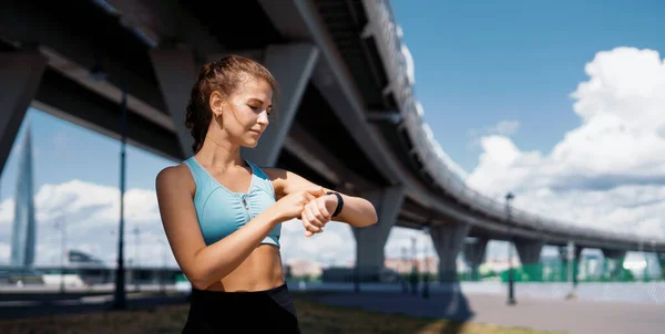 Antrenör vücudu germek için egzersiz yapar. Sokakta spor yapan bir kadın. Fitness ve sağlıklı bir yaşam tarzı. Ellerindeki nabzı ölçmek için akıllı bir saat..