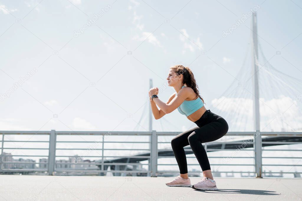 A strong female fitness trainer does strength exercises squats outdoors. Healthy lifestyle concept. Slim figure and comfortable shoes.