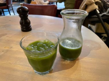 Bir bardak ve cam sürahi, içinde süper yeşil meyve suyu olan bir restoranın masasına oturmuş. Buzlar bardaklarda. Sağlık içeceği.