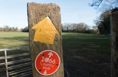 Sarı bir ok ve '1066 kır yürüyüşü' yazan yuvarlak bir tabela. Tahta bir postanın kırmızı arka planında beyaz harf. Doğu Sussex.UK