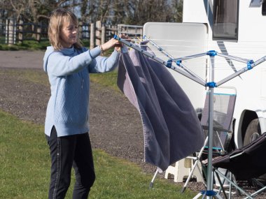 Bir bayan karavan sahibi arka planda eğlence aracıyla havlusunu döner bir saç kurutma makinesine asar. Güneşli bir gün.