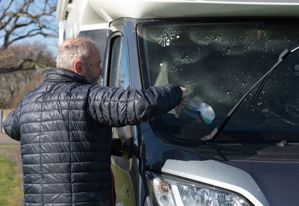 Sakallı bir erkek karavan sahibi ön camını mavi sıvıyla temizliyor. Güneşli bir gün.