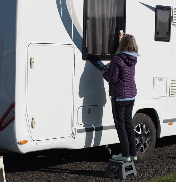 A lady motorhome owner cleans her recreational vehicle window.She stands on a step to reach the window.RV