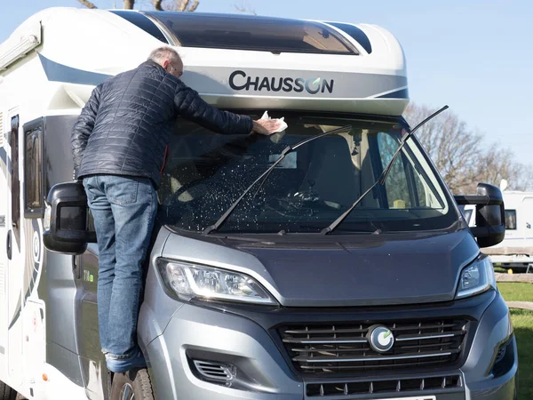 Erkek bir karavan sahibi ön camını temizler. Erişmek için lastiğin üzerinde durur. Güneşli bir gün. Chausson.