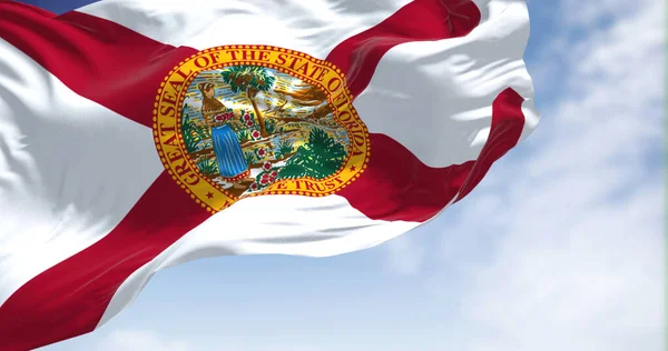 The Florida state flag waving in the wind. In the background there is a clear sky. Florida is a state located in the Southeastern region of the United States