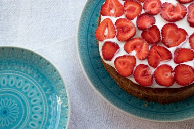 Çilekli kek ve mavi tabakta krema. Beyaz masa örtüsü üzerinde Yaz Çileği ve Kremalı Kek. Üst görünüm, boşluğu kopyala. Yaz ortası yemeği.