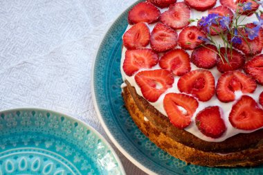 Çilekli kek ve mavi tabakta krema. Beyaz masa örtüsü üzerinde Yaz Çileği ve Kremalı Kek. Üst görünüm, boşluğu kopyala. Yaz ortası yemeği.