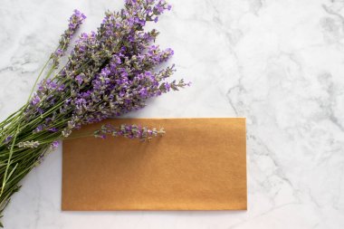 Yaz kırtasiyesi, lavanta çiçekleri ve beyaz mermer arka planda boş kraft kağıdı zarflı düğün davetiyesi ile hayat dolu. Davetiye kartının kopyasını çıkar. Üst görünüm, düz görünüm, kopyalama alanı