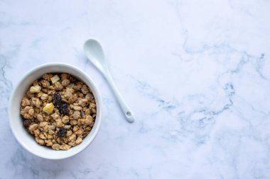 Bowl of homemade granola cereal with ceramic spoon on white marble table background. Top view, flat lay, copy space. Healthy eating concept.