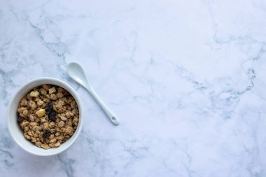 Bowl of homemade granola cereal with ceramic spoon on white marble table background. Top view, flat lay, copy space. Healthy eating concept.