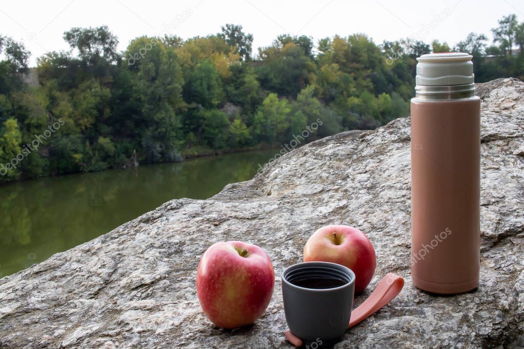 Beige thermos bottle, cup of tea or coffee, and red apple on rock over the river. Beautiful summer, spring, autumn landscape. Picnic on the rock