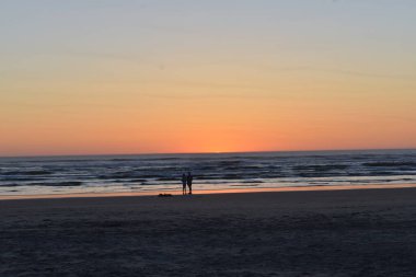 Akşam gökyüzünde gün batımı, kumsalda bir çift manzaranın tadını çıkarıyor. 