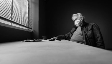 Evde tek başına oturan, maske takan ve gazete okuyan yalnız bir son sınıf öğrencisi. Virüs vakası acil durum önlemleri ve yaşlı güvenlik kavramı.