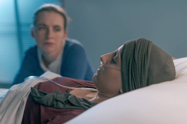 Hastanede genç arkadaşına kanser konusunda yardım eden ilgili bir kadın, yatağının yanında oturuyor ve onu kontrol ediyor.