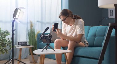 Vlogger evde video çekiyor, cep telefonunu gösteriyor ve gülümsüyor.