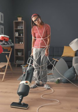 Evde ev işleri yapan hayal kırıklığına uğramış ev hanımı: Kablolara sarılmış, yerleri süpürüyor ve dağınık bir odayı temizliyor