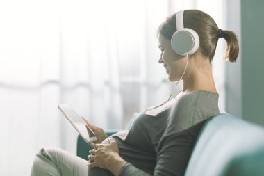 Evdeki koltukta oturan ve dijital tabletine bağlanan gülümseyen hamile kadın, kulaklık takıyor ve internetten video oynuyor.