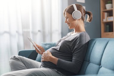 Evdeki koltukta oturan ve dijital tabletine bağlanan gülümseyen hamile kadın, kulaklık takıyor ve internetten video oynuyor.