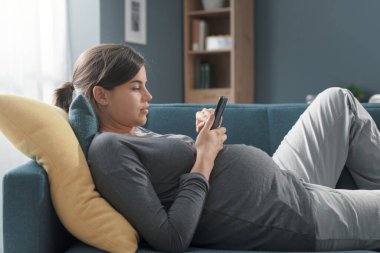 Hamile bir kadın kanepeye uzanıp telefonuyla iletişim kuruyor. Rahatlıyor ve sosyal iletişim kuruyor.