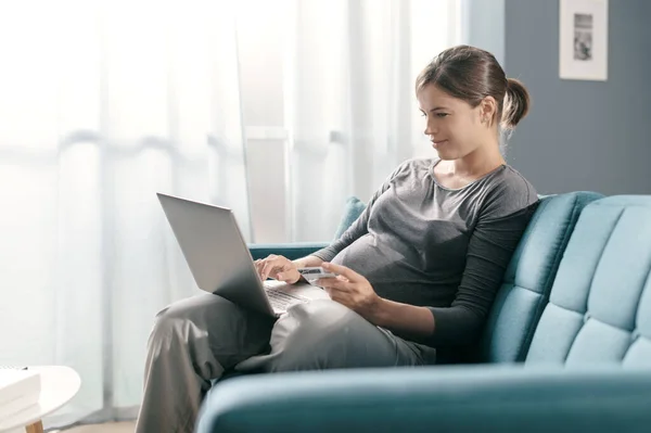 怀孕妇女坐在沙发上 在家网上购物 她正在用笔记本电脑连接 并使用信用卡 — 图库照片