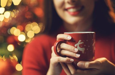 Mutlu genç bir kadın Noel 'i evde kutluyor, elinde bir fincan sıcak çay, gülümsüyor, süslenmiş bir ağaç ve arka planda ışıklar tutuyor.