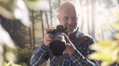 Ormanda yürüyüp çekim yapan profesyonel fotoğrafçı kameraya gülümsüyor.