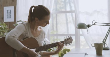 Evdeki masada oturan genç kadın gitar çalıyor ve müzik, yaratıcılık ve müzik konsepti oluşturuyor.