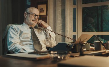 Klasik tarzda bir gazeteci masada oturuyor ve telefonla konuşuyor.