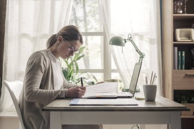 Genç iş kadını masada oturuyor ve evde çalışıyor. Bilgisayarıyla bağlantı kuruyor.