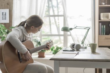 Evde bir masanın yanında oturan ve gitar çalan genç bir kadın.
