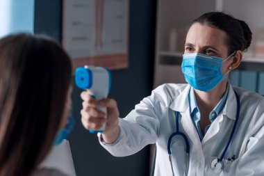 Cerrahi maskeli profesyonel bir doktor. Hastanın ateşini kızılötesi alın termometresiyle ölçüyor.