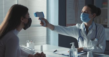 Cerrahi maskeli profesyonel bir doktor. Hastanın ateşini kızılötesi alın termometresiyle ölçüyor.