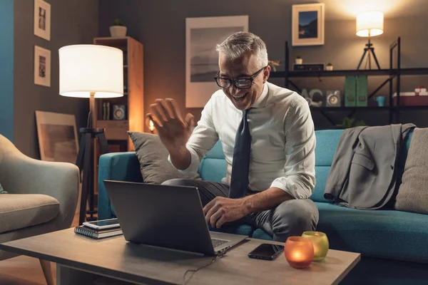 自宅のソファに座ってノートパソコンと接続している企業のビジネスマン 彼は誰かをビデオ通話し コンピュータのウェブカメラで手を振っています — ストック写真