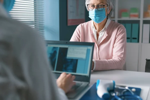 医者はオフィスで顔のマスクを持つ高齢者を満たし ラップトップ上の電子医療記録を確認します — ストック写真